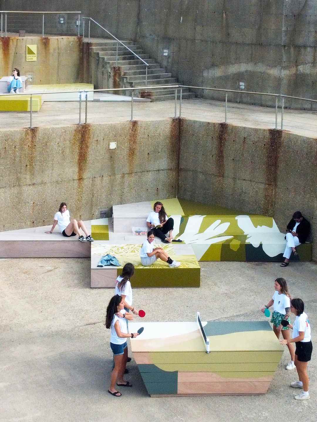 Visitatori seduti sulle piattaforme in legno dipinte della Discesa Mare all'Argentiera, con un tavolo da ping pong